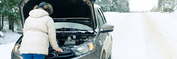 A person in a puffy white coat stands in front of the open hood of a car on a snowy road.